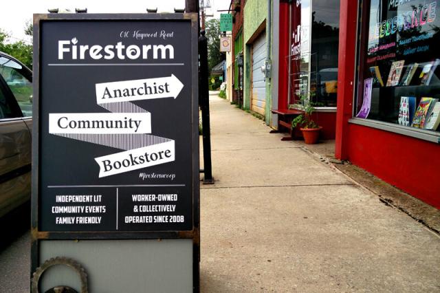 A photo of the sidewalk in front of Firestorm. Across from the red building is an a-frame sign that reads "Anarchist Community Bookstore" with smaller text reading "independent lit, community events, family friendly" and "worker-owned & collective operated since 2008".
