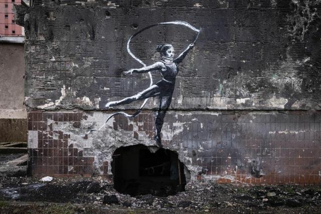Photograph of a Banksy stencil artwork that depicts a black and white dancer waving a long ribbon on a stick. The piece is painted on the side of a building damaged by bombing in Ukraine.