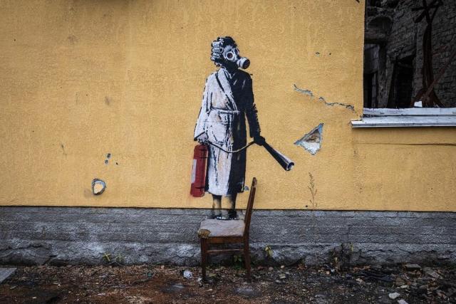 Photograph of a Banksy stencil artwork that depicts a woman in hair curlers and gas mask holding a red fire extinguisher. She appears to be standing on a chair in front of the stencil artwork that is painted on the facade of a damaged building in Ukraine. 