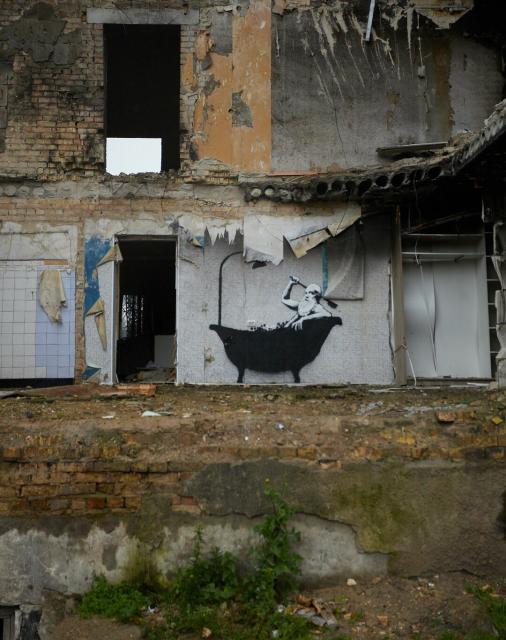 Photograph of a Banksy stencil artwork that depicts a black and white bearded man in a bathtub scrubbing his back. The work is painted on the wall of an actual bathroom that has been opened up due to bombing. 