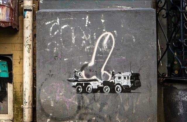 Photograph of a Banksy stencil artwork that depicts a military truck and soldiers who are moving a phallic graffiti tag. 