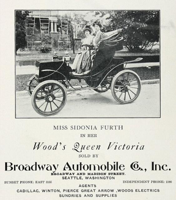 Miss Sidonia Furth and another woman in Seattle Washington in a Wood's  Queen Victoria electric automobile. They are driving the vehicle on a road near a large home in the background. Both women are well dressed, appear to be upper class (and would need to be to afford an electric car of this quality) and have elaborate feathered hats. 