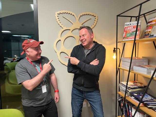 Kevin in a red hat and grey t shirt and Toby in a black hoodie.They are stood in front of a Raspberry Pi logo on the wall behind them and are facing each other pointing and laughing.