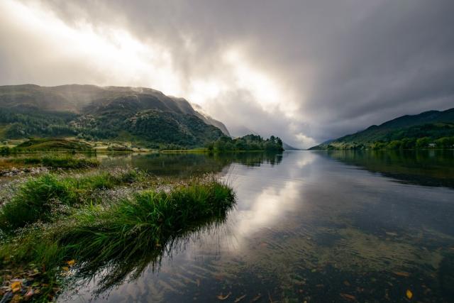 Picture taken from the shore of a lake with hills in the background and a dramatic light from the sun going through clouds. 