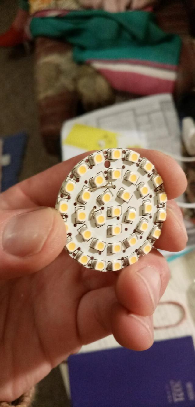 A photograph of an array of tiny LED bulbs set in a circle. This is the inside of an LED lightbulb. There are 36 tiny circular bulbs on a circular breadboard being held by a hand. The board is around five centimetres in diameter.