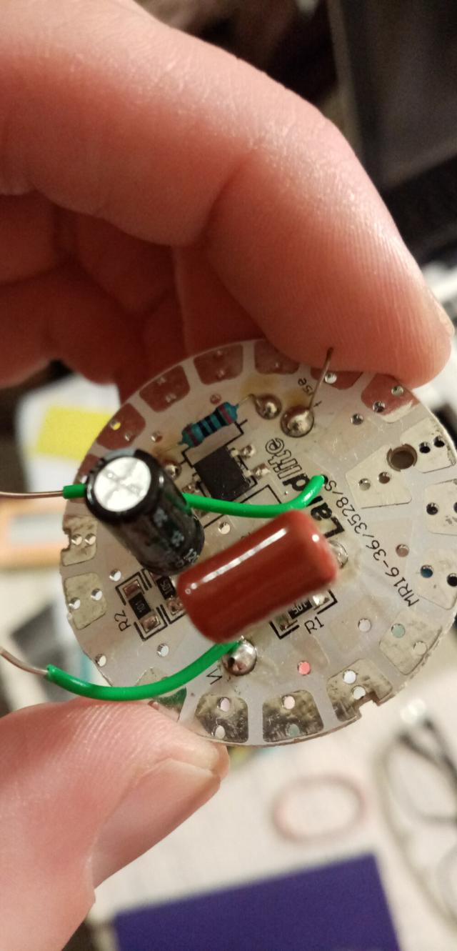 The reverse of the circuit board from inside an LED lightbulb. There is a resistor (blue resistor with the following stripes: brown, black, black, black, red), a transistor and a couple of other modules that I don't recognise. It is being held by a hand.