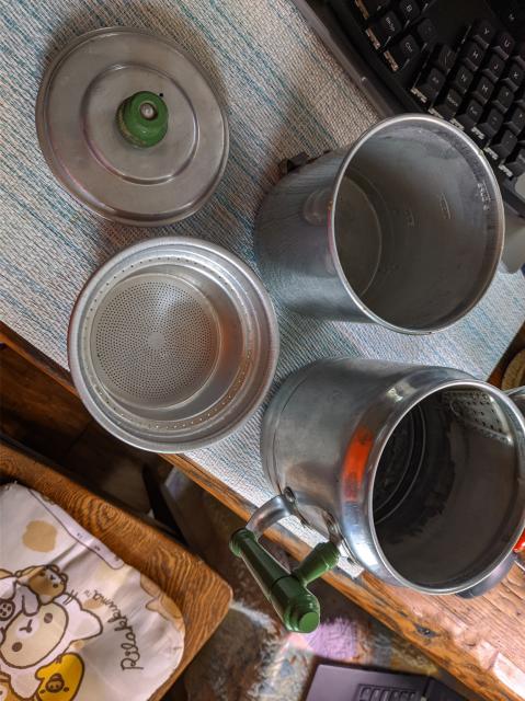 Four pieces of the aluminum coffee pot - a decanter base, a flat-bottomed strainer basket filter, a top dripper cylinder with tiny holes in the bottom, and a lid. 