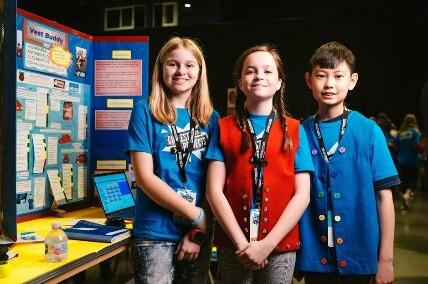 Three young people stood in front of a school project wearing blue tee shirts and lanyards round their necks
