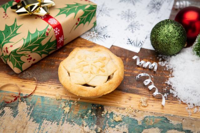 A mince pie on a wooden table with the corner of a wrapped present visible, some baubles, and fake snow. The mince pie has a snowflake pressed into the pastry on the lid.