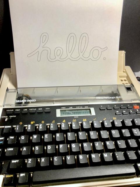 A Silver Reed EB50 typewriter plotter with paper in the platen that says hello in a lower case cursive outline font.