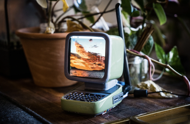 A retro looking light green PC with a steam punky aesthetic on a wooden table with houseplants around it. Onthe screen of the PC is a seascape with orange clouds and a blue sky.