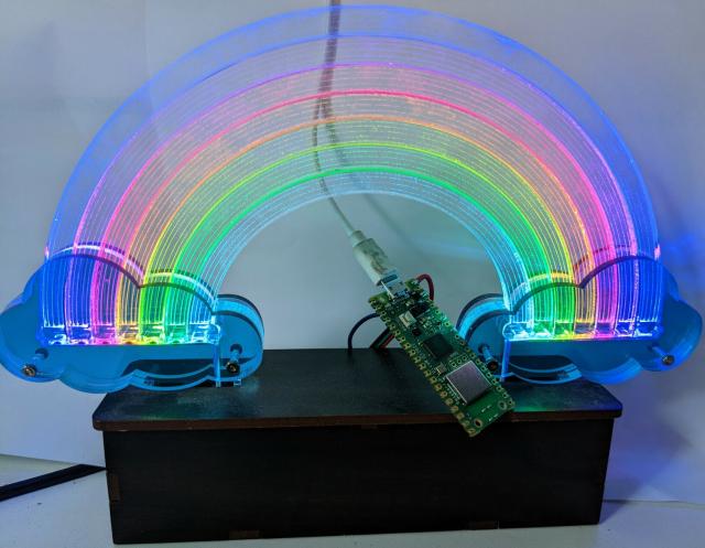 An image of a laser cut rainbow made from 7 semicircles of clear acrylic with translucent blue clouds are each end of the rainbow. Each of the rainbow's arcs is illuminated at either end by an addressable LED, and the acrylic rainbow and clouds are mounted on top of a laser cut wooden box. In the middle of a rainbow is a Raspberry Pi Pico W microcontroller with a USB cable connected and hookup wires leading from the Pico to control the LEDs illuminating the rainbow.