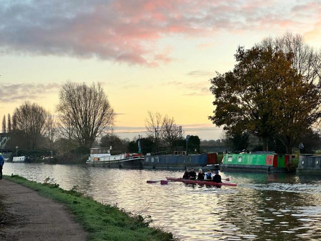 A view of the River Cam with a dirt path on the left, with rowers on the river, and boats along the left side. On the far side of the river is a bunch of bare trees and sunrise in a partially cloudy sky.