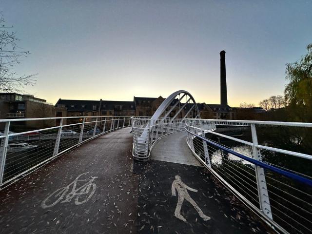 A view of the surface of a bridge which splits into two paths, on the left for bicycles and on the right for pedestrians. In the background is a row of buildings and a smokestack.