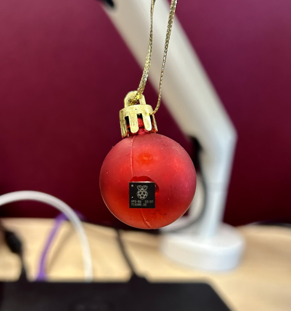 A mini red Christmas bauble hanging from the bottom of a desktop monitor. The bauble has an RP2040 chip glued to it