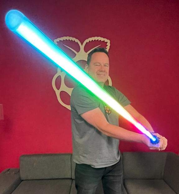 Toby stood in front of a red wall with a Raspberry Pi logo behind him holding a rainbow coloured lightsaber towards the camera