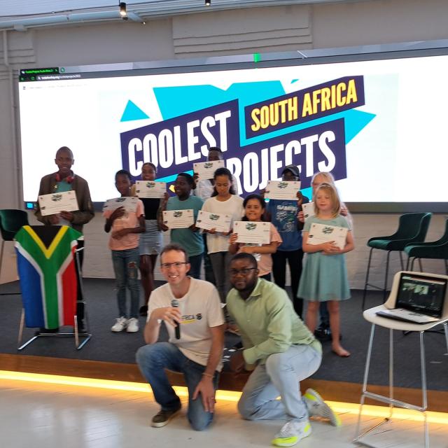 Group of 7-17 year olds proudly holding up certificates and grinning. The coolest projects south africa logo is behind them a south african flag to the left, and a chromebook with remote participants on a stand to the right. Mentors crouch on the floor in front. 