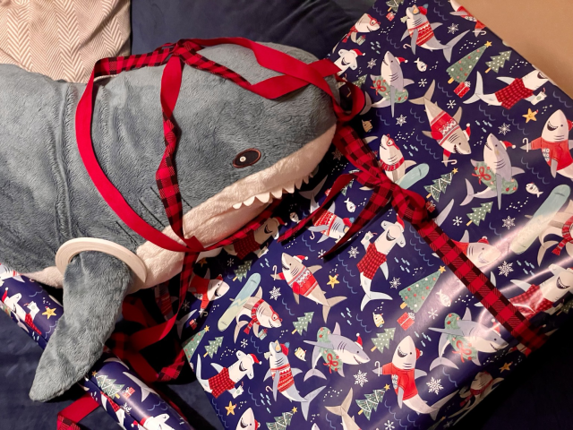 A large IKEA shark is covered in gift wrapping ribbon next to a large gift wrapped in gift wrapping paper with sharks on it
