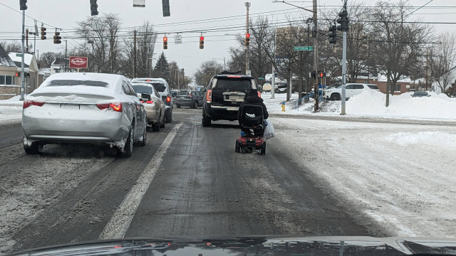 A stroad with lots of cars, and one person in an electric wheelchair, because all the snow makes it impossible to use the sidewalks.