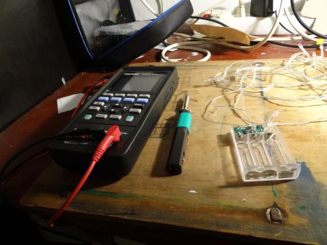 A digital multimeter next to an unplugged portable soldering iron and an LED light chain with its transparent plastic box open, showing the PCB and the battery compartment.