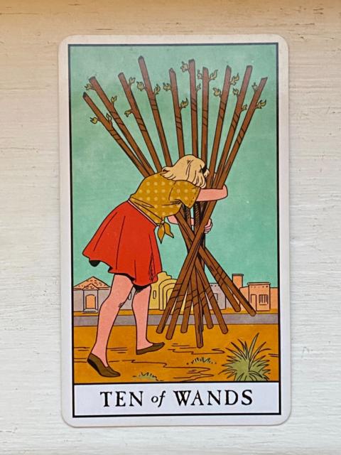 Ten of Wands from the Modern Witch Tarot: a white woman with blonde hair, a yellow dotted top, and a red skirt stumbles toward a desert town, bent over with the effort of holding the loose bundle of tall staffs she holds vertically in front of her