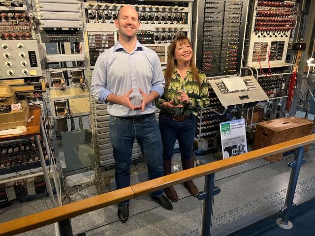 Liz and Eben Upton holding small glass awards stood in front of the enigma code breaking machine. From the Raspberry Pi co-founders honoured by The National Museum of Computing article. 