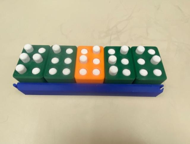 Five green and orange blocks with poppable white dots spell out the word "raise" on a deep blue rack. Each block is 3d printed, about an inch tall and just over half an inch wide. 