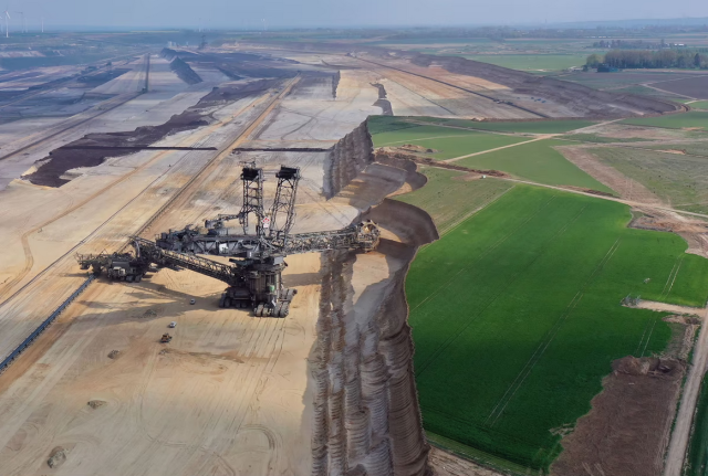 An aerial view of the Tagebau Hambach coal mine in Germany, and the equipment used to carve into the earth.  The Excavator 288, a mobile mining rig, is gargantuan, approximately a hundred meters tall and 200 meters long.  The mine itself is kilometers long and wide.  The floor of the mine is below the undisturbed ground level by more than half the rig’s height.  Outside the mine is a green plain of former farmland, and inside the mine is a dusty plain of exposed dirt.  It is truly difficult to convey the scale of the scene in words.