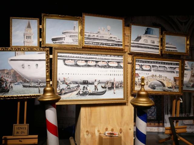 Some gold frames containing paintings showing gondoliers in the foreground and a huge cruise ship in the background. It is suffocating the city of Venice.