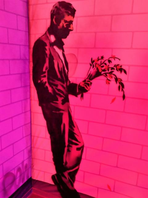 Image of a man leaning against a wall. He is holding a bunch of flowers with their heads hanging slightly.