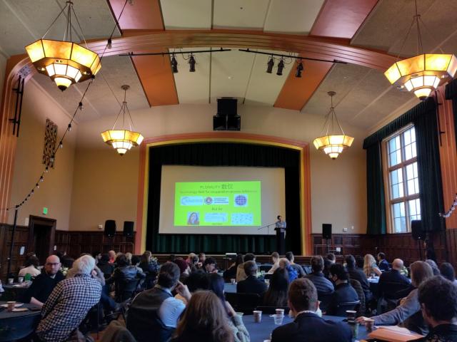 Lecture hall with a yellow slide on the screen that says on top "PLURALITY"