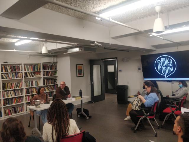 Dr. Pierre sits at a table with cohost behind there is a well-stocked bookshelf, a screen to the right displays The People's Forum logo. The crowd is fully engaged with the seminar.