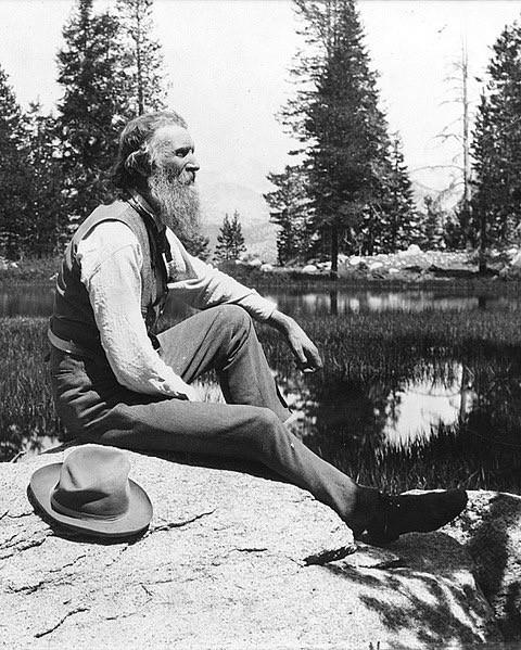 John Muir, full-length portrait, facing right, seated on rock with lake and trees in background, circa 1902. Library of Congress.