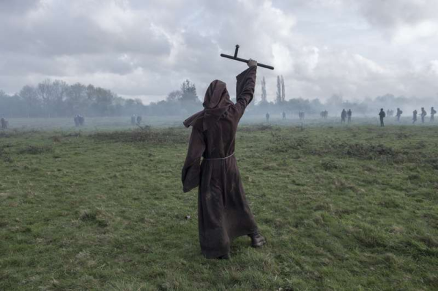 A photograph of a demonstrator at the ZAD ("Zone to Defend") at Notre-Dame-des-Landes, facing off with riot police during their attempt to evict many of the residents in 2018. Clad in a brown cowl belted with a length of rope, he holds aloft a police baton as if it were a crucifix.