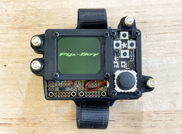 Black Pip Boy on a wooden table. Green screen says "Pip Boy". Connectors and wires are visible. 