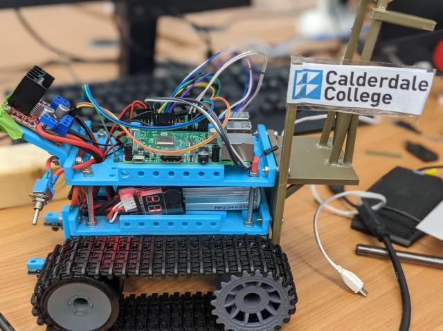 A small tracked robot, the upper sections are 3D printed in blue filament and it has a small flag with the "Calderdale College" logo on it. 