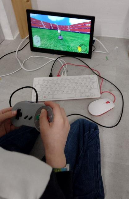 SuperTuxKart wird auf einem RaspberryPi 400 gespielt, sichtbar sind die Hände eines Kindes, dass ein Gamepad in den Händen hält.