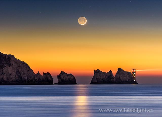 The crescent moon, looking more like a full moon due to earthshine perfectly above the Needles. The sun has just gone down on a cold day, giving the last colour of the day. The Needles lighthouse is illuminated and the sea calm. Colours are orange, yellows, blues and purples.