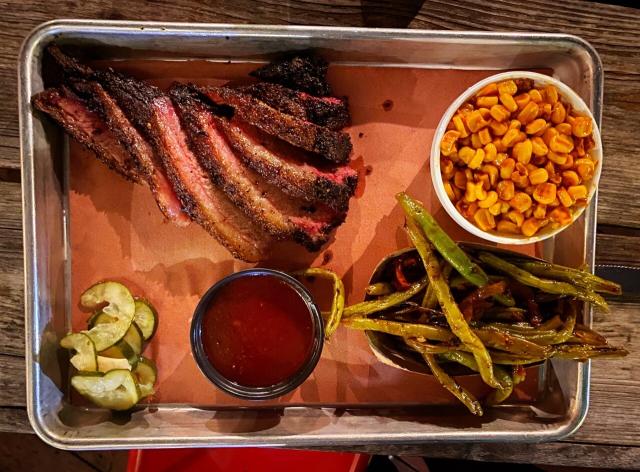 Metal tray holding a barbecue meal of sliced brisket, corn, green beans, barbecue sauce, and a small pile of pickles.