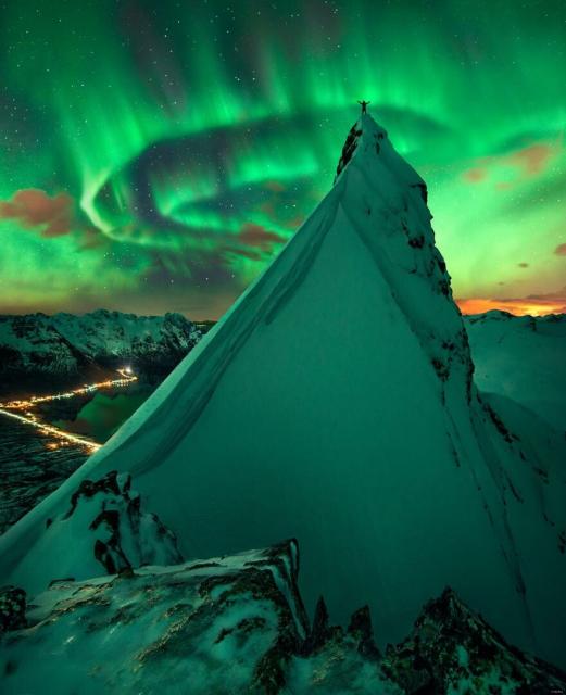 A person stands on a steep snow-covered hill with their arms raised. In the distance green aurora are visible. Past that stars are visible.