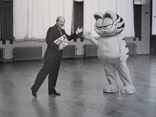 Jim Davis and someone wearing a Garfield suit in a large, empty room. Davis is smiling and gesturing towards Garfield. Garfield is doing something approximating a wave. 