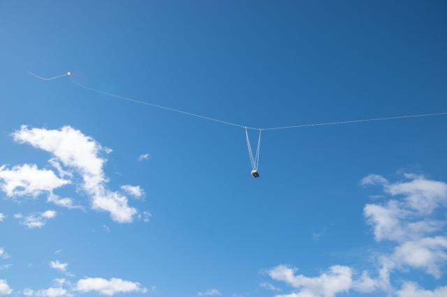 Sobre un cielo azul una cuerda sostiene enganchada la cámara y al fondo a la izquierda se ve la cometa al final de la línea