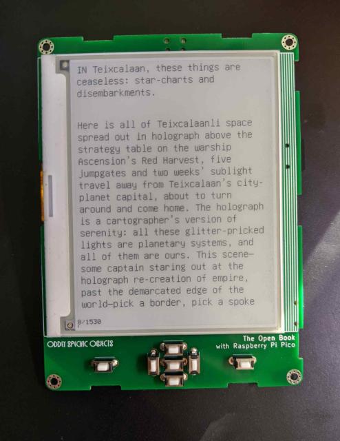 Small ebook front panel - an e-ink screen (displaying the first page of Arkady Martine's A Memory Called Empire) on a green circuit board, with seven buttons below the screen. The circuit board reads "ODDLY SPECIFIC OBJECTS" and "The Open Book with Raspberry Pi Pico"