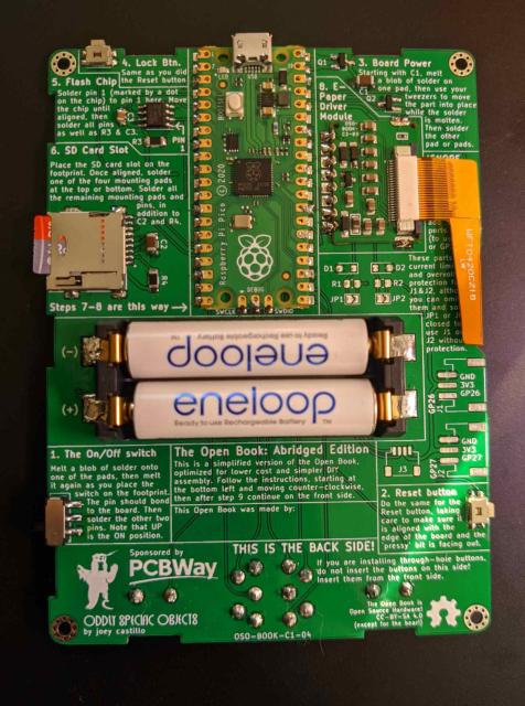Back of the open book circuit board, showing a fair bit of clumsy surface-mount soldering. Raspberry pi pico at top center, above two AAA batteries.