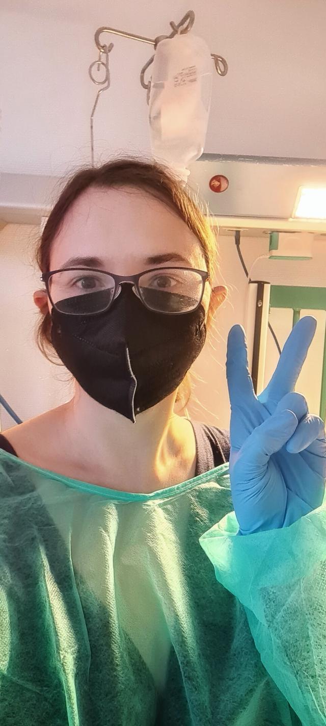 White woman with brown hair in a ponytail. She's wearing glasses, a green hospital robe, blue gloves, and a black mask. There is an IV bag, bed part, and a light in the background. 