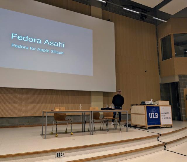 Presenter on stage at the FOSDEM conference. Large text on screen says "Fedora Asahi — Fedora on Asahi Silicon"