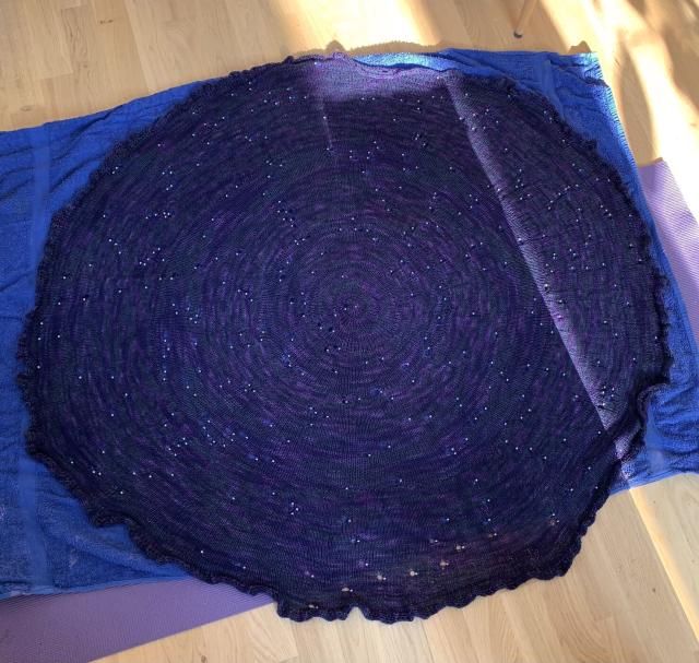 A purple circular shawl on a blue towel on a rubber mat. The shawl has beads placed to represent the stars on the northern hemisphere, with the northern star at the center. 