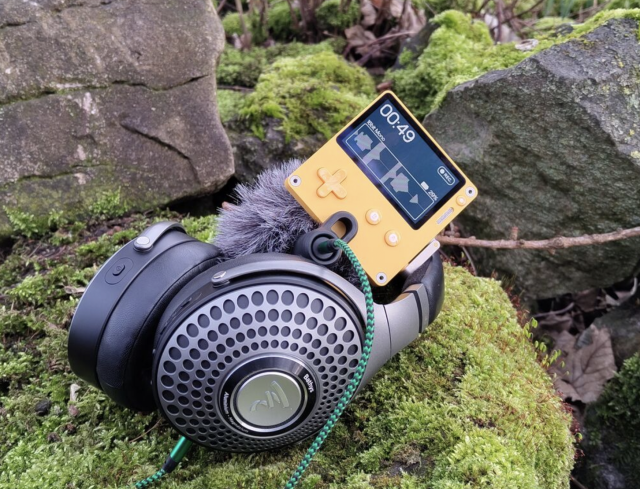 A Playdate resting on a pair of Focal Bathys headphones, the Playdate has a Rode microphone attached which has a 'dead cat' wind muffler. The whole ensemble is resing on a moss covered rock in Cumbria