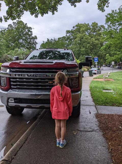 Photo of an 8 year old girl dressed in hot pink, standing in front of the grill of a red Chevy Silverado parked on a sidewalk. The top of the hood is several inches above the girls head.