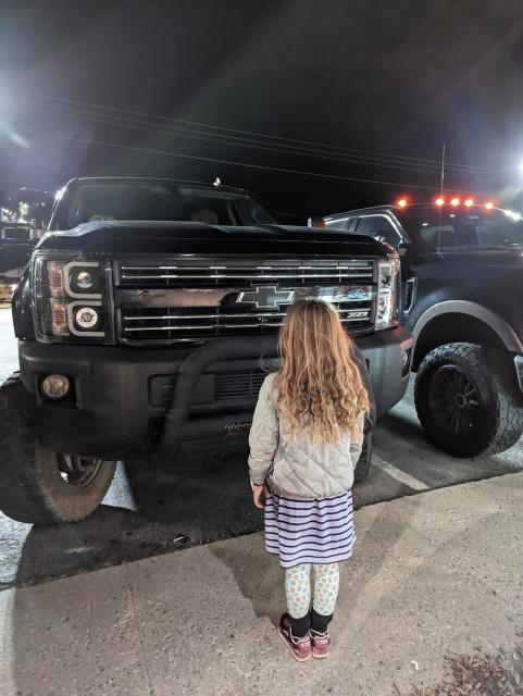 A photo of a nine year old girl, in a striped dress, standing in front of the grill of a black Chevy Silverado. The top of the hood on this truck is even higher above the girl's head, despite her recent growth spurt.